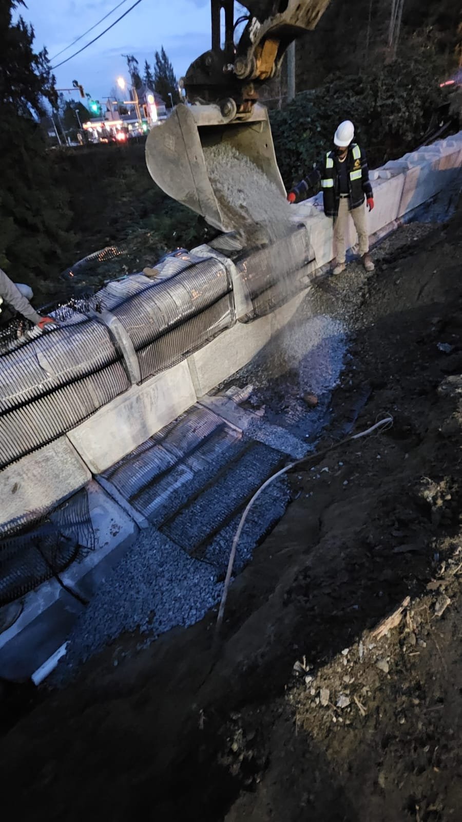 Excavator working on retaining wall backfill at dusk